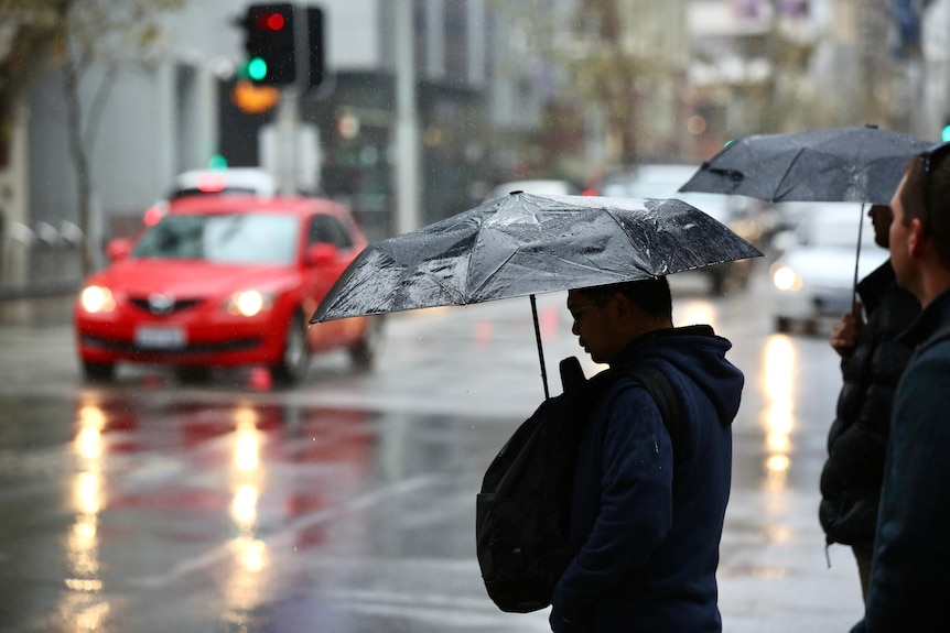 A man silhouetted against the wet road and traffic stands with an umbrella in the rain wearing a backpack on his front