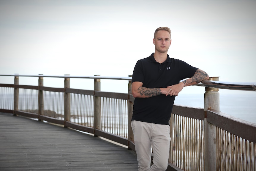 A man leans on a silver rail on a beach-side timber boardwalk with the ocean in the background.