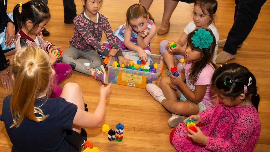 A group of young children gather around a box full of colourful toys on the floor