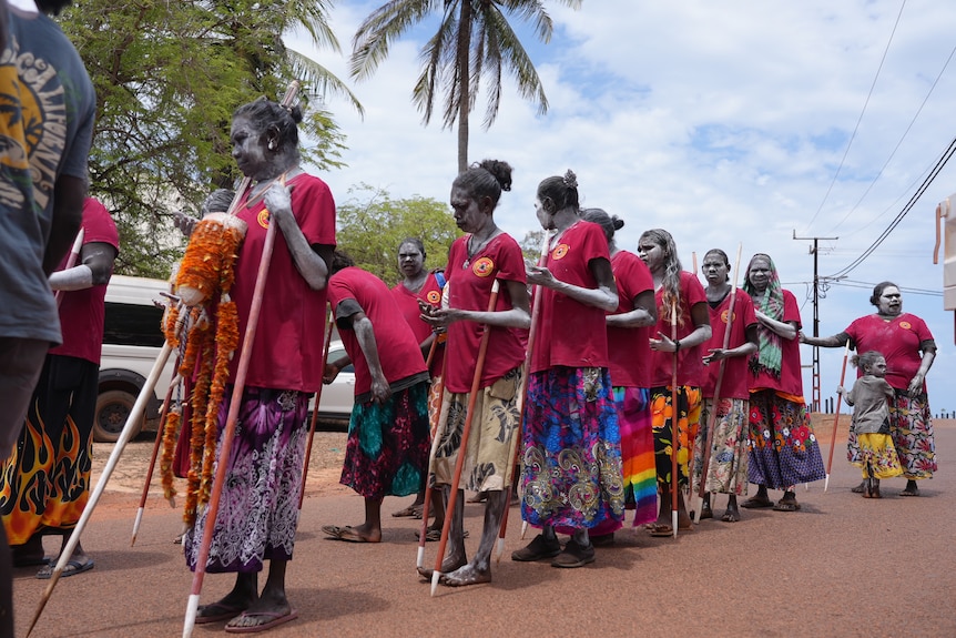 Group of Indigenous women in red shirts hold sticks and stand in a line with traditional paint on