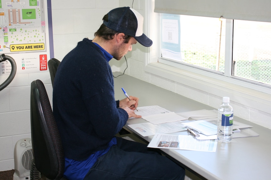 a man in a cap and black jumper sits at a desk filling out forms. 