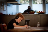 A child is writing on a notepad and wearing headphones while studying from home.