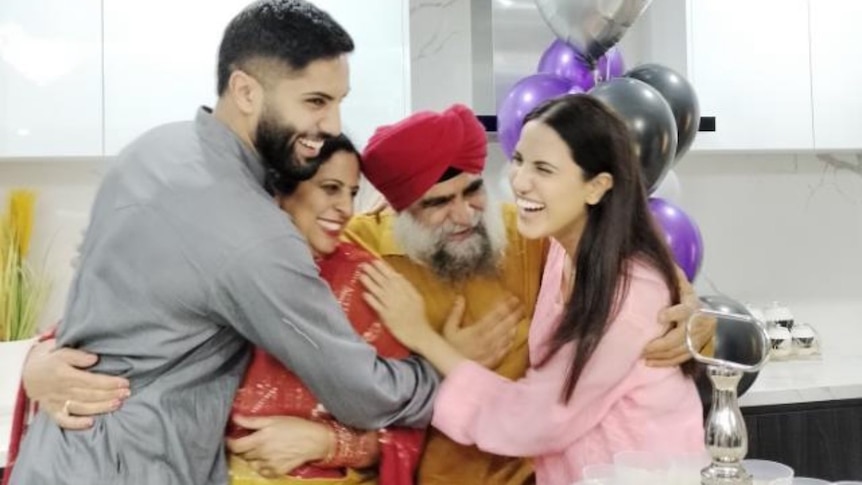 A young Sikh woman hugs  her dad who wears a red turban, and her mum and brother