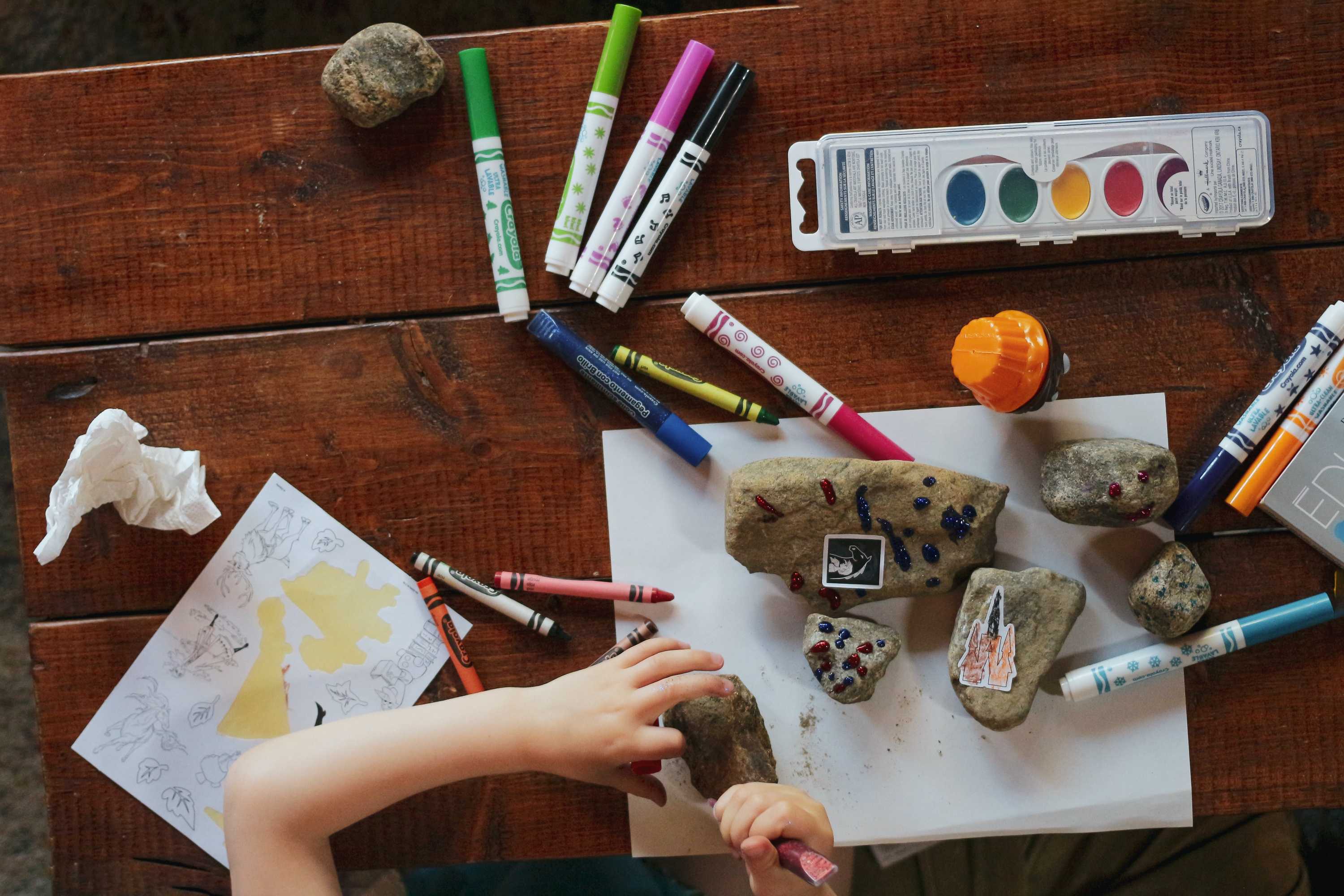An aerial view of a table with pens and paints, and a child's hand making craft with rocks