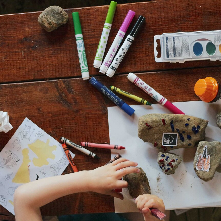 An aerial view of a table with pens and paints, and a child's hand making craft with rocks