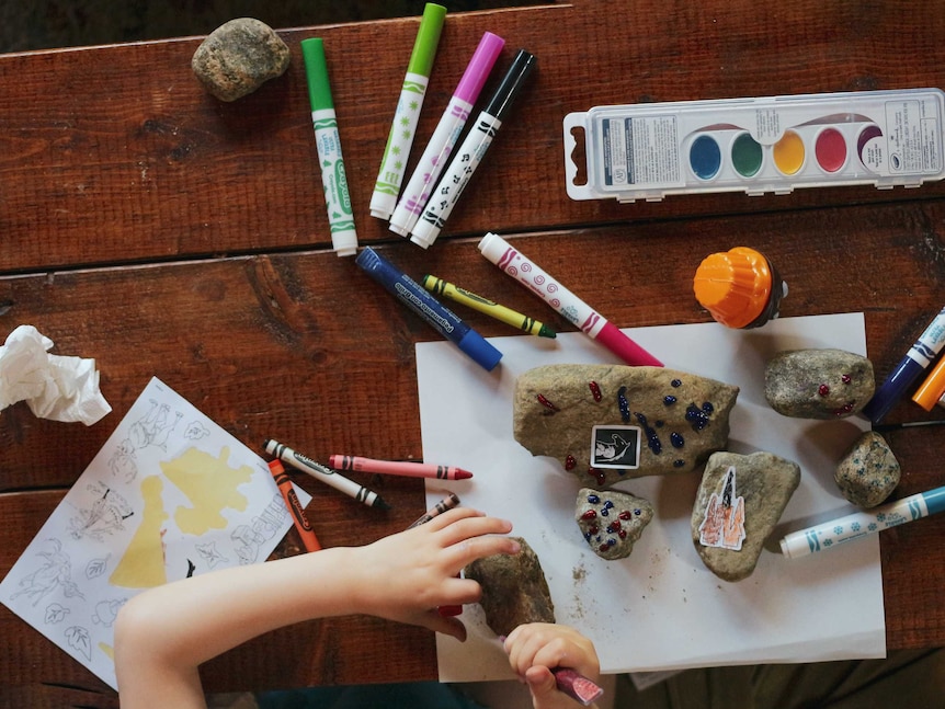 An aerial view of a table with pens and paints, and a child's hand making craft with rocks