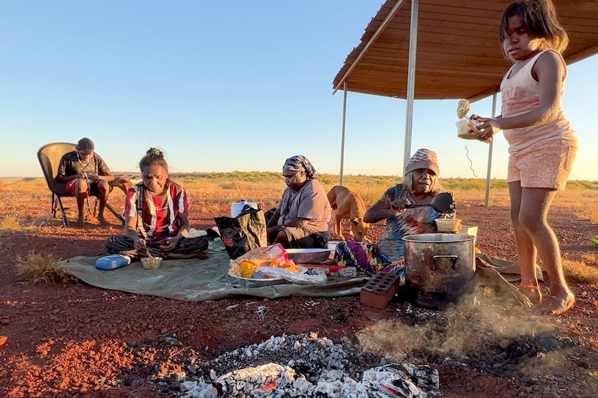 An Indigenous woman serves dinner around a campfire. 
