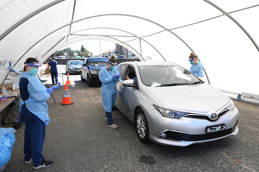 Three nurses wearing protective gowns and facial equipment standing around a car