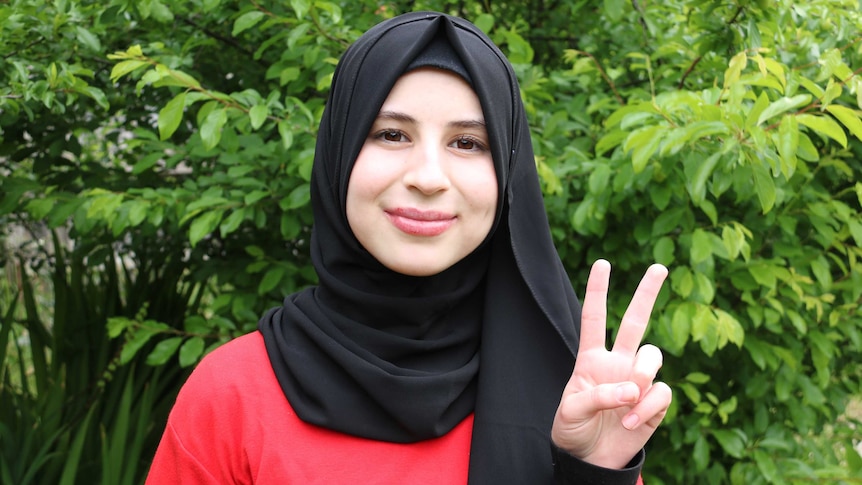 A photo of Nour smiling and raises her index and middle finger showing a sign of peace