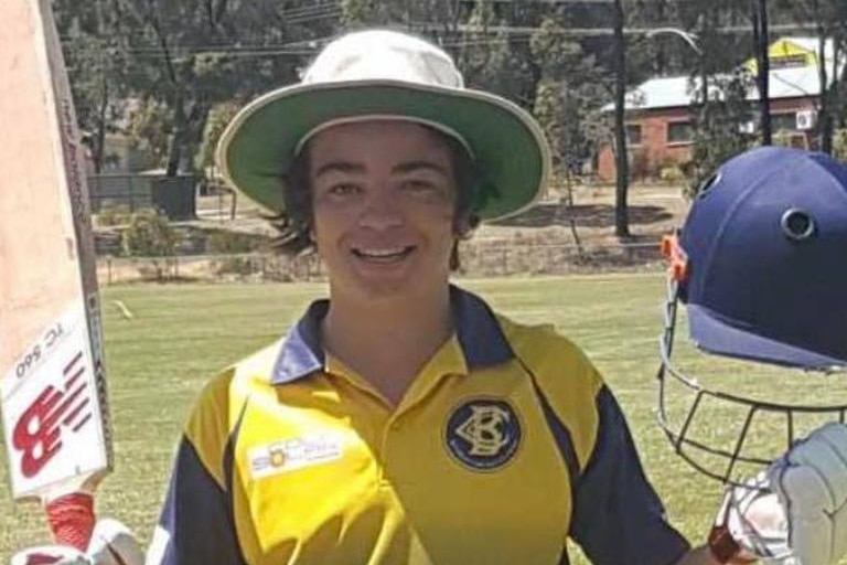 teenage boy in cricket gear, holding bat and cap, with hat on