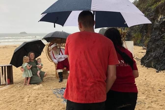 A photographer and assistance stand under an umbrella taking photos of Santa on the beach with a family in the rain.