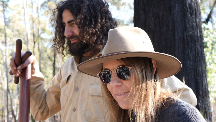 Indigenous man holding clapsticks in background, woman in hat and sunglasses in foreground