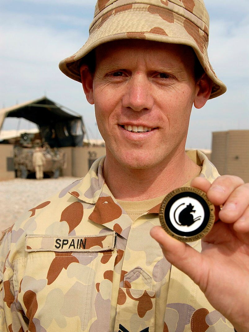 A smiling man in soldier's uniform stands under a blue sky, holds up a large coin with a black mouse on it.