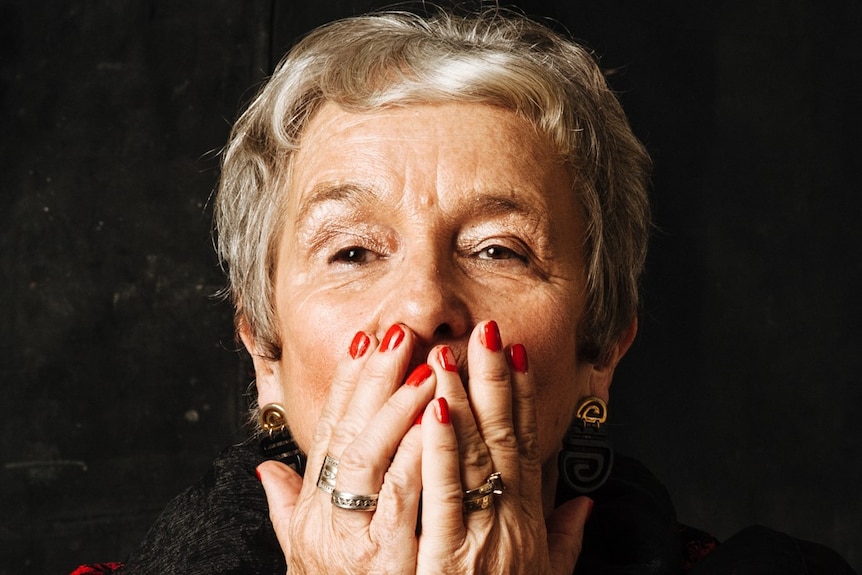 Older woman with short hair looking at the camera, hands clasped over mouth, cheeky eyes.