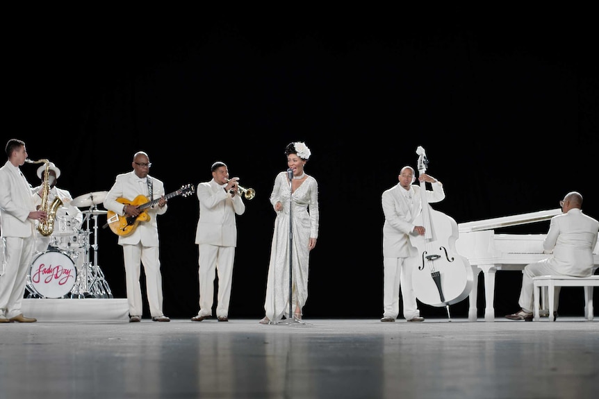 Billie Holiday, dressed in white, sings with her band, also in white, playing instruments