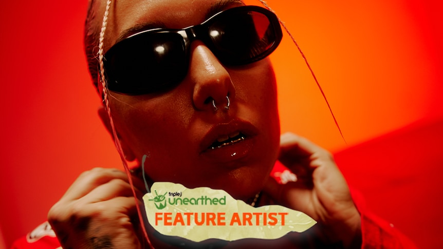 Sophiya, wearing black sunglasses and a nose ring, faces the camera in front of a red background