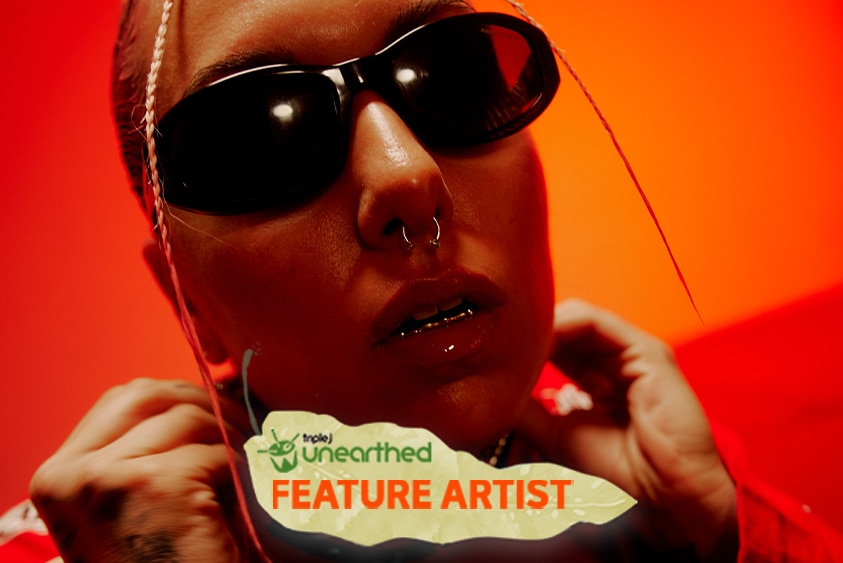 Sophiya, wearing black sunglasses and a nose ring, faces the camera in front of a red background