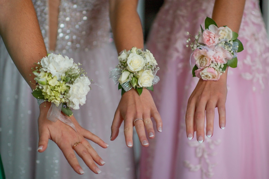 Three women in ball dresses extend their wrists to show off their floral corsages