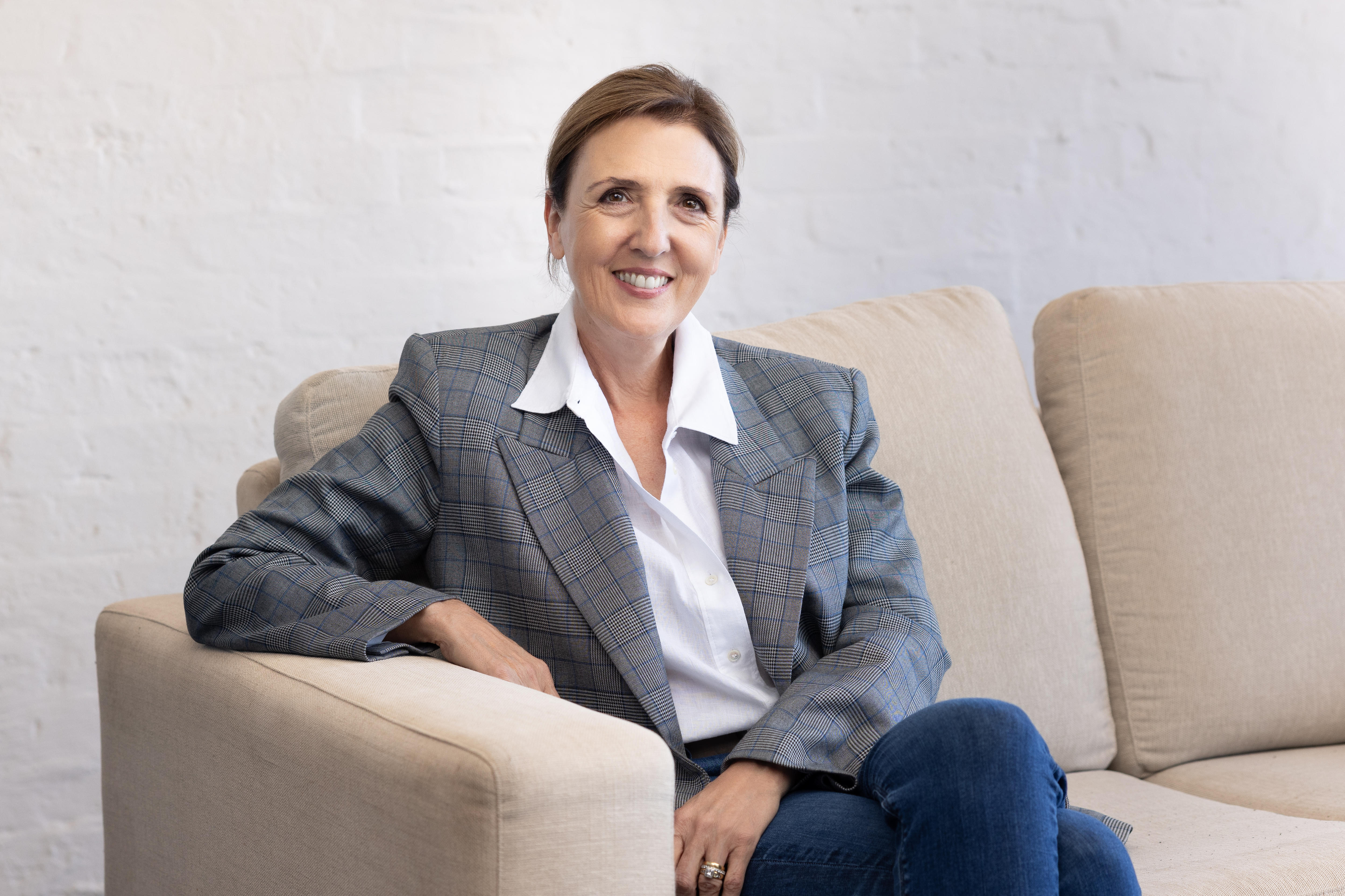 A fair-skinned brunette woman, Tawnya, in a grey blazer and white shirt sits smiling on a cream couch.
