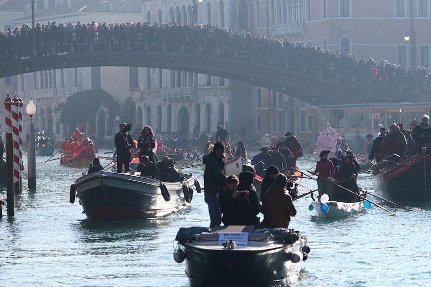 Gondolas pass under a packed bridge on a busy canal
