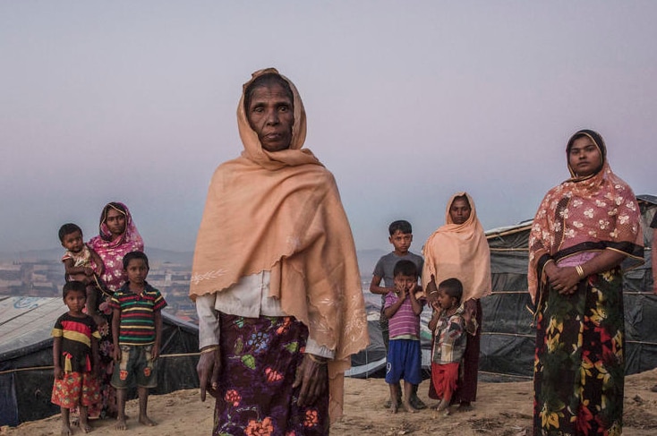 A elderly woman stands surrounded by younger family members amid basic shelters.