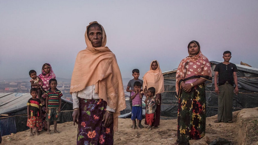 A elderly woman stands surrounded by younger family members amid basic shelters.