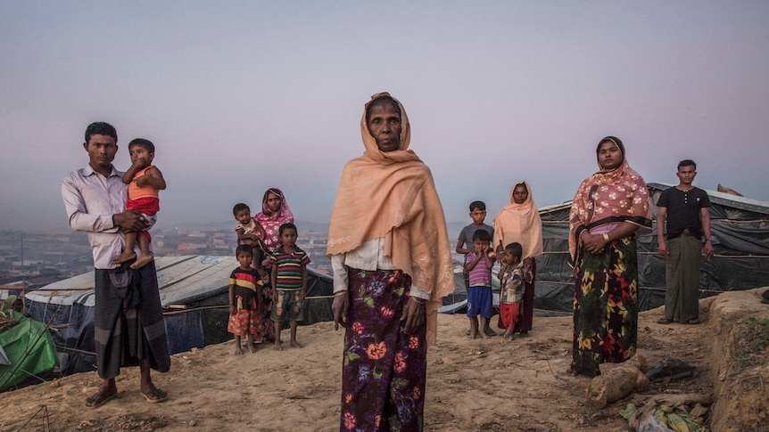 A elderly woman stands surrounded by younger family members amid basic shelters.