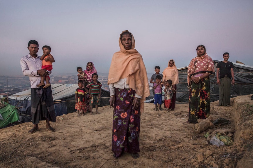 A elderly woman stands surrounded by younger family members amid basic shelters.