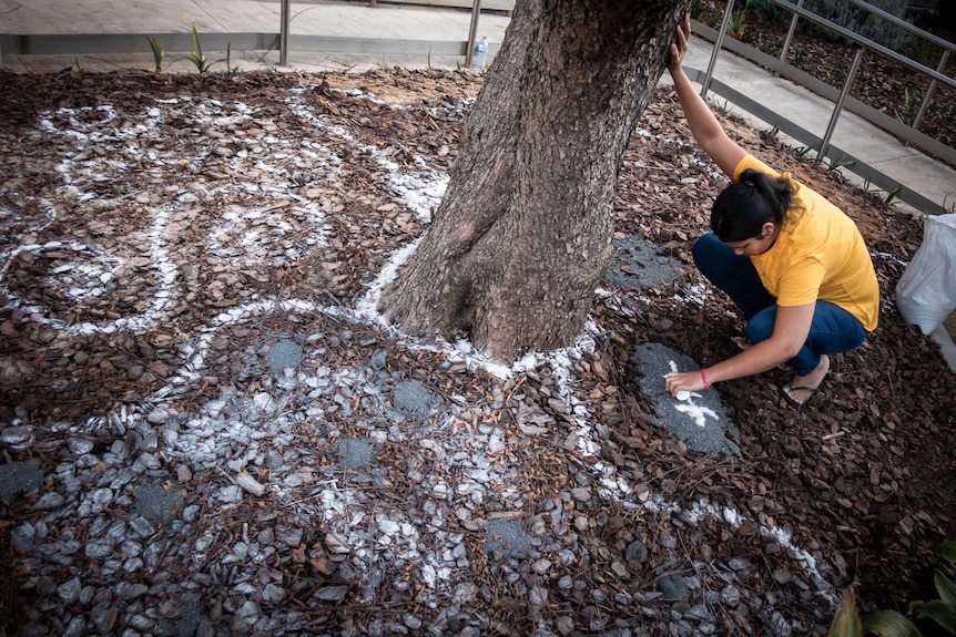 At the base of a tree, someone pours sand on the ground, adding to a mural.