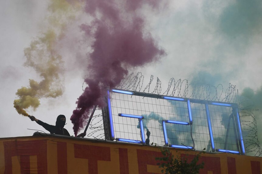 A balaclava-clad protester holds a flare in each hand billowing yellow and purpose smoke next to an LED G20 sign