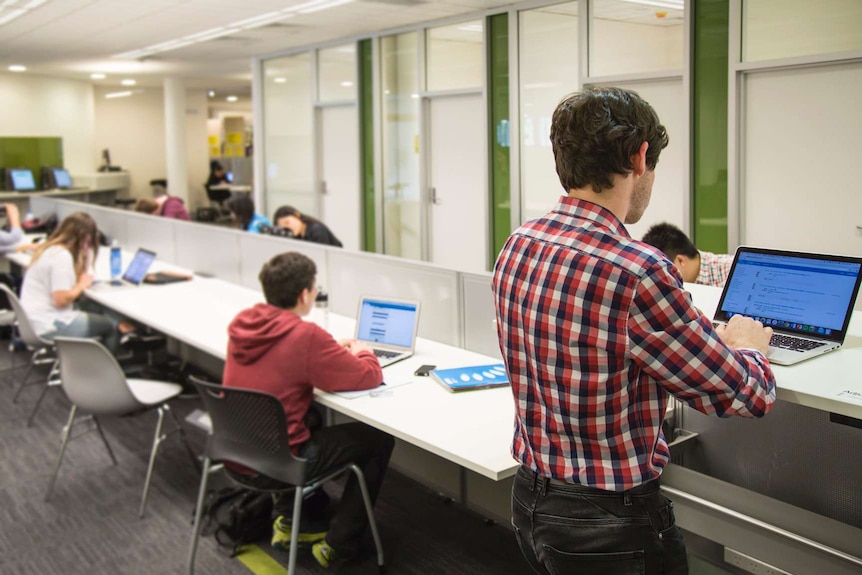 Students sit and stand on adjustable desks at a university library.