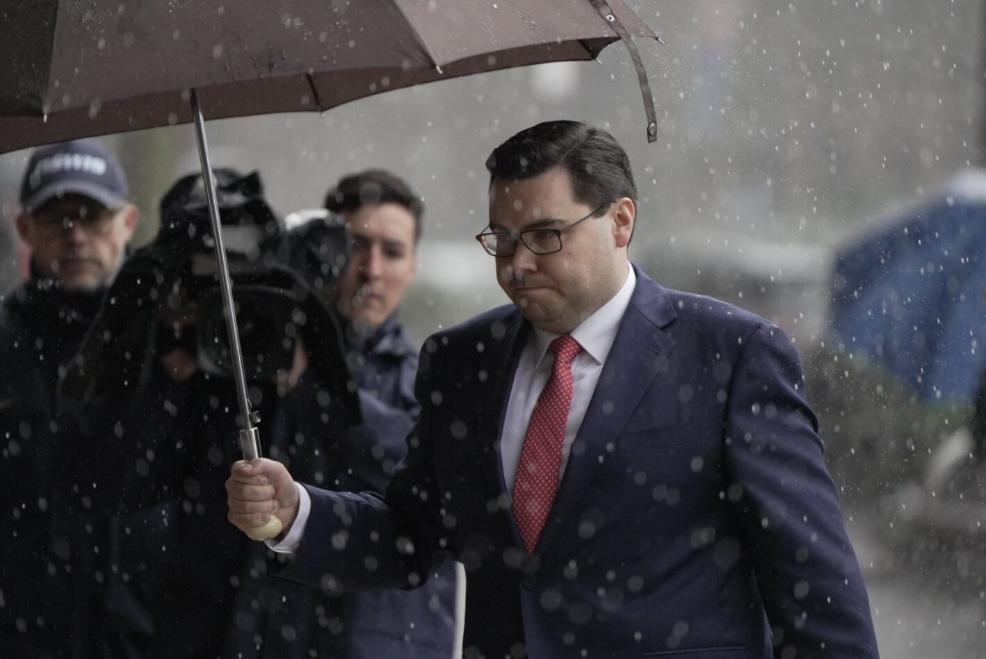A man with glasses in a suit holds an umbrella.