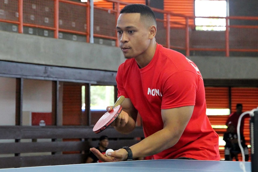 A powerfully built Tongan man in a red shirt prepares to serve during a game of table tennis.