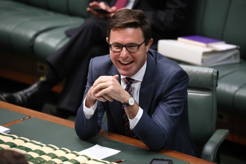 Littleproud has his elbows on a table, and is laughing. He has dark-rimmed glasses.