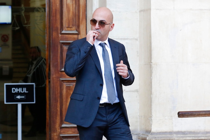 A bald man in a grey pinstripe suit places a cigarette in his mouth and walks in front of a wooden doorway.