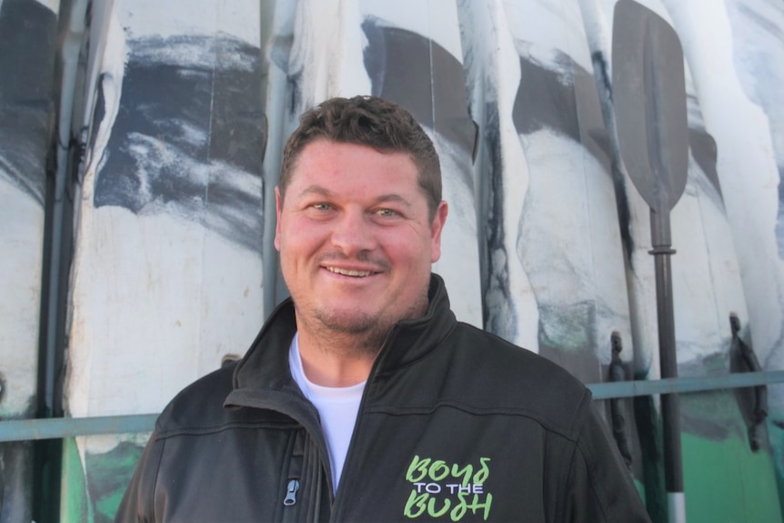 A man  in a black jumper stands in front of a row of up right canoes 