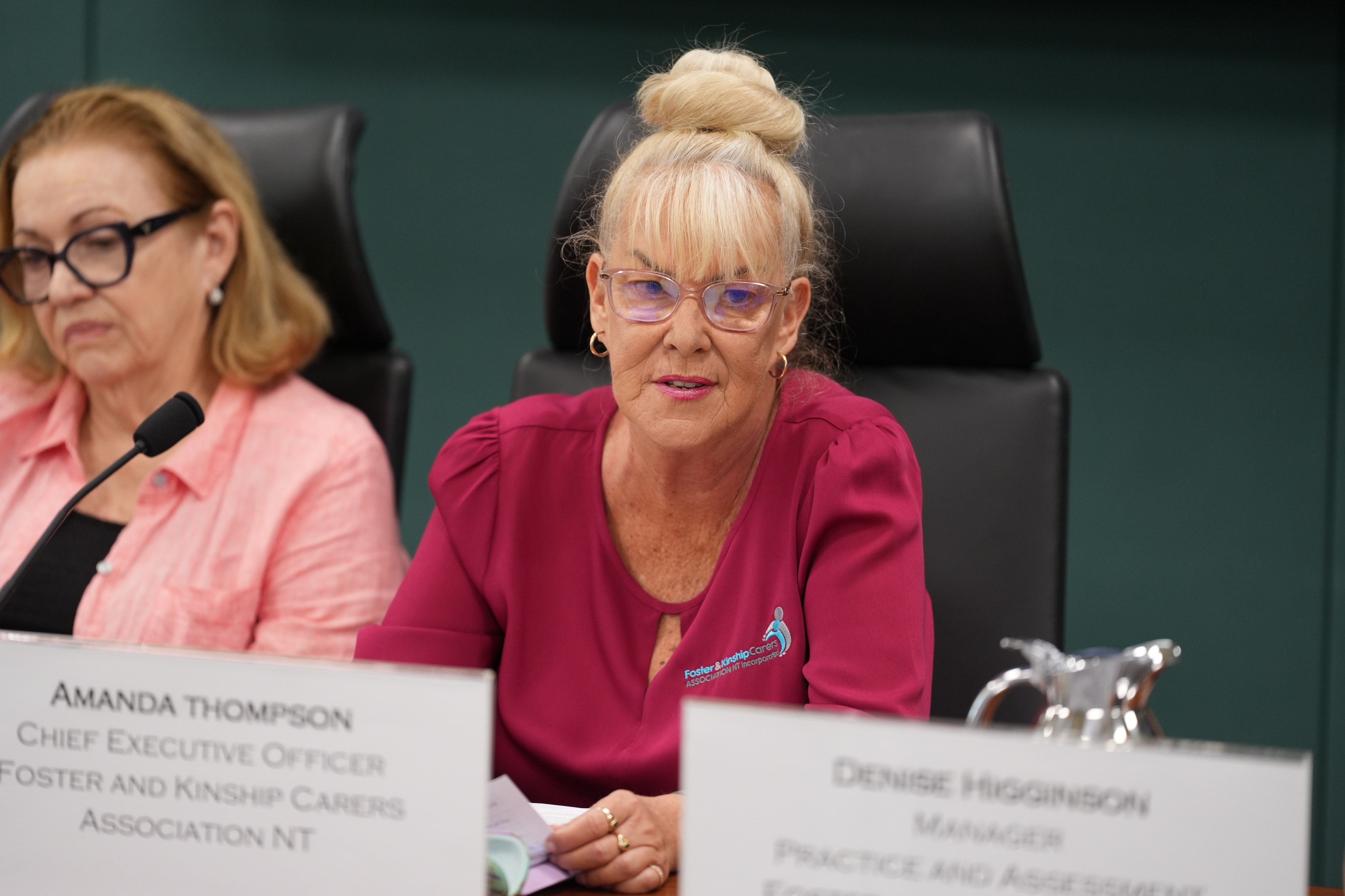 A blonde woman in a pink blouse wearing glasses as she speaks into a microphone.