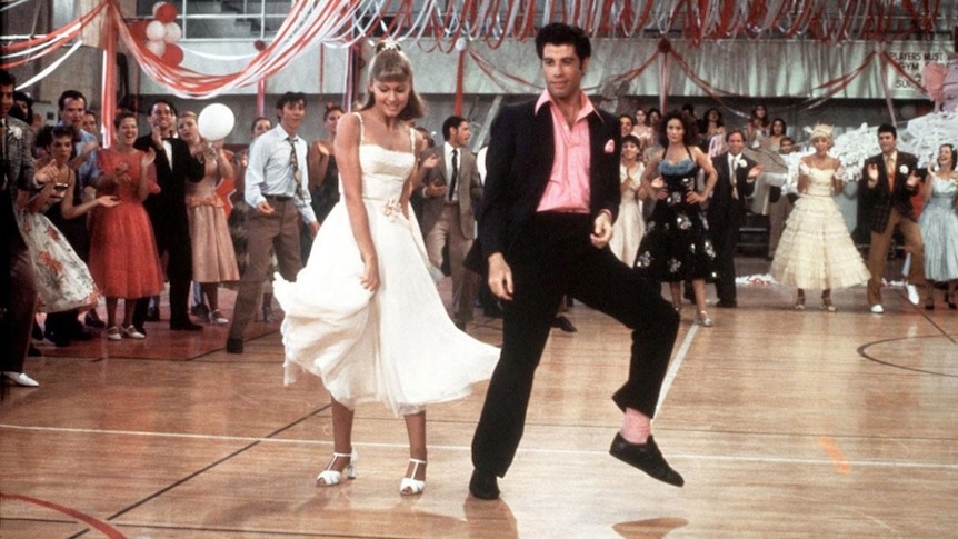 Two people in 1950s ballroom attire dancing on a dance floor while people watch in the background 