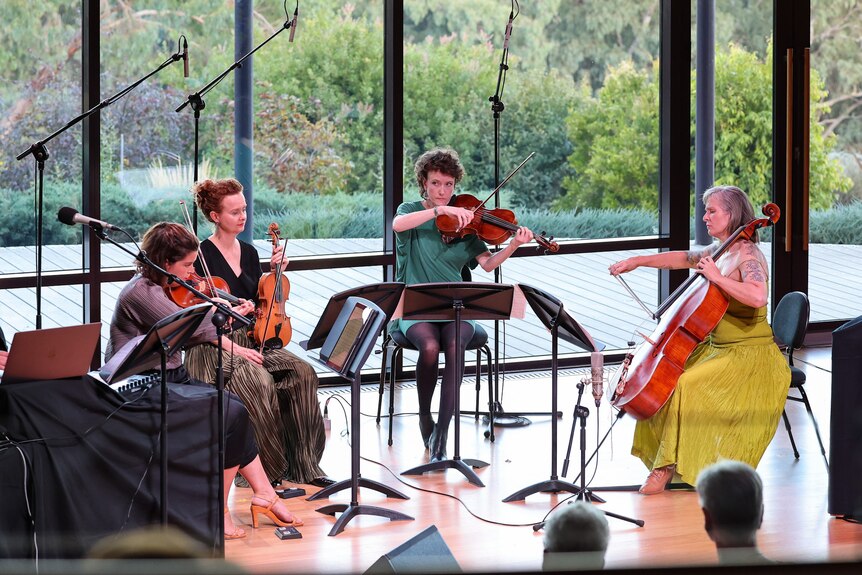 String quartet seated on stage, playing, with gardens seen through glass behind them.