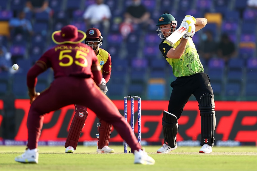 An Australian batter punches a shot through the field on the off-side as West Indies fielders watch.