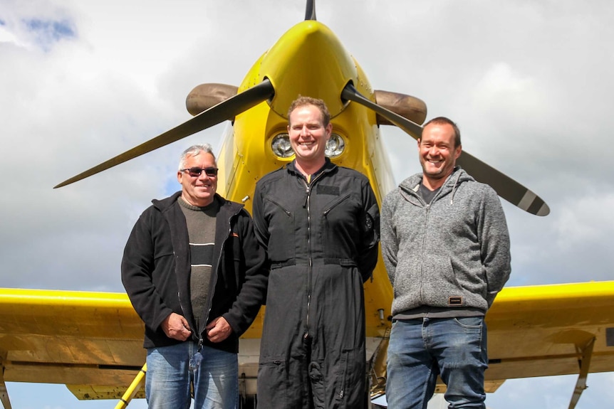 Three smiling men stand in front of the nose of an aerial seeding plane
