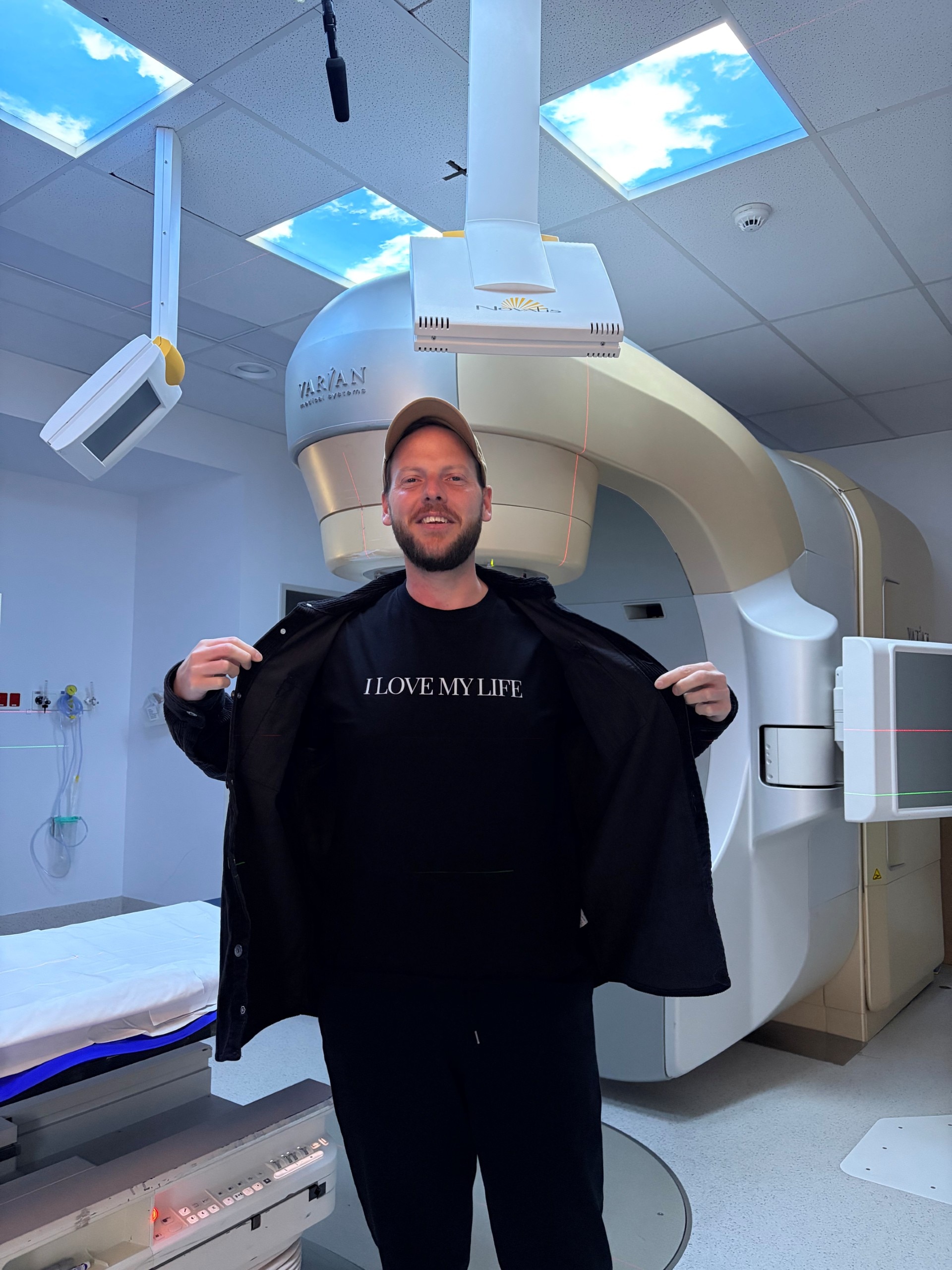 A young man with a cap in a hospital setting wears a black shirt that says i love life. 