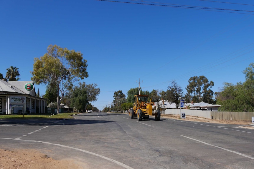 A country town streetscape with a pub on the left and a road grader driving past on the right.