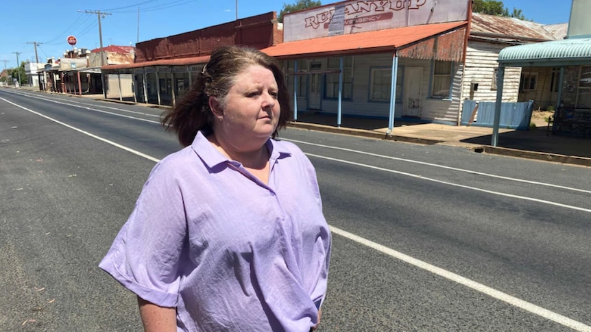 A middle-agd woman stares stoically down an abandoned main street. The sun is shining brightly.