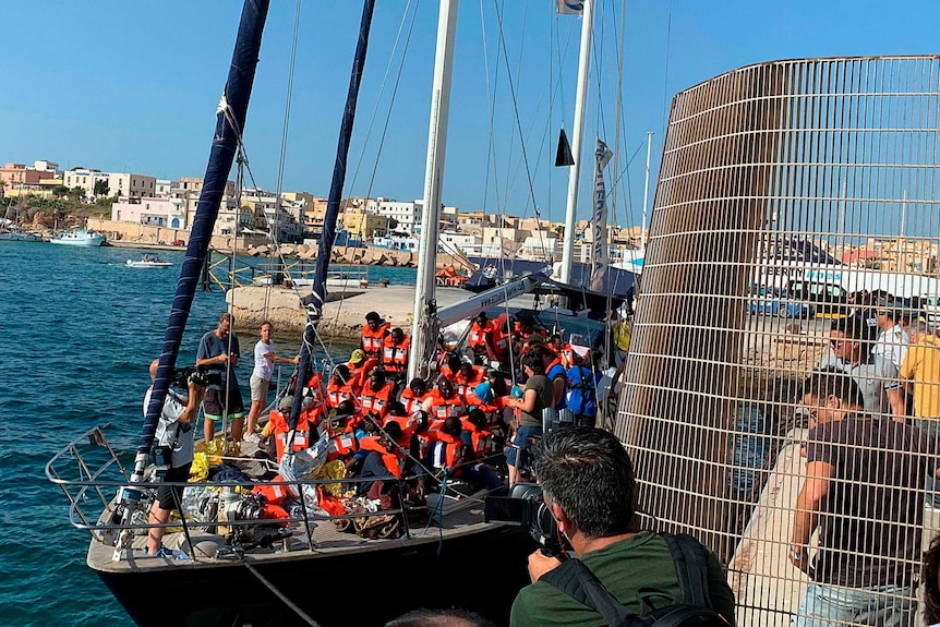 A group of men people in lifejackets fill the deck of a boat as it approaches a fenced jetty.