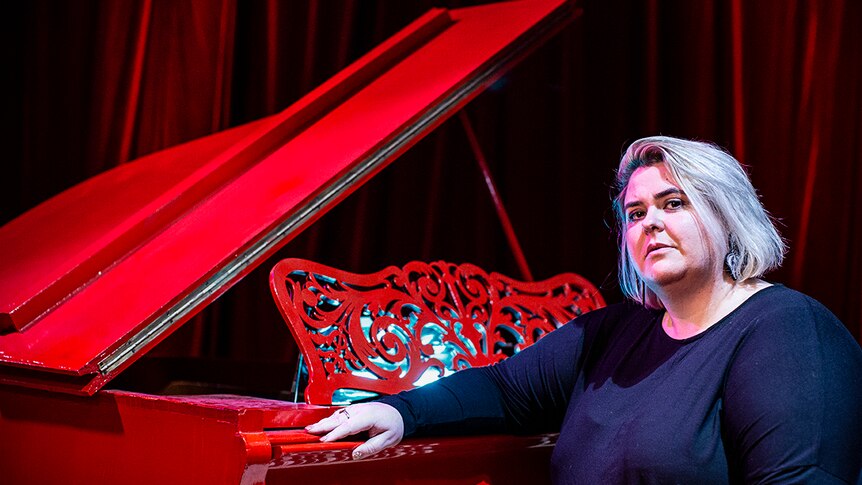 Colour photo of Danielle Harvey sitting by a blood red piano inside a set from immersive theatre production A Midnight Visit.