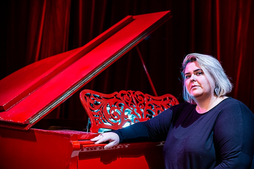 Colour photo of Danielle Harvey sitting by a blood red piano inside a set from immersive theatre production A Midnight Visit.