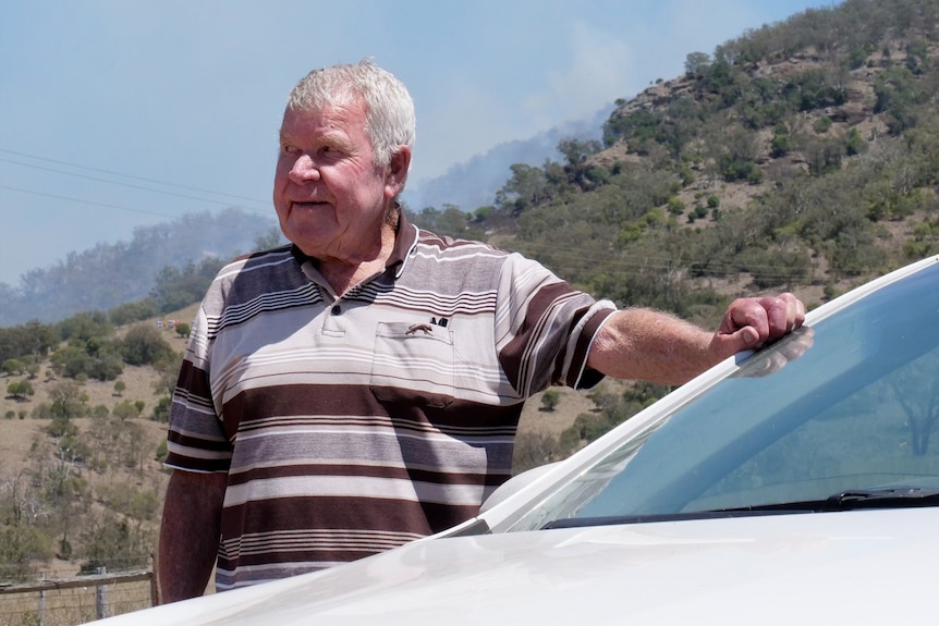 An elderly man stands near the bonnet of a car, looking to the side of the camera.
