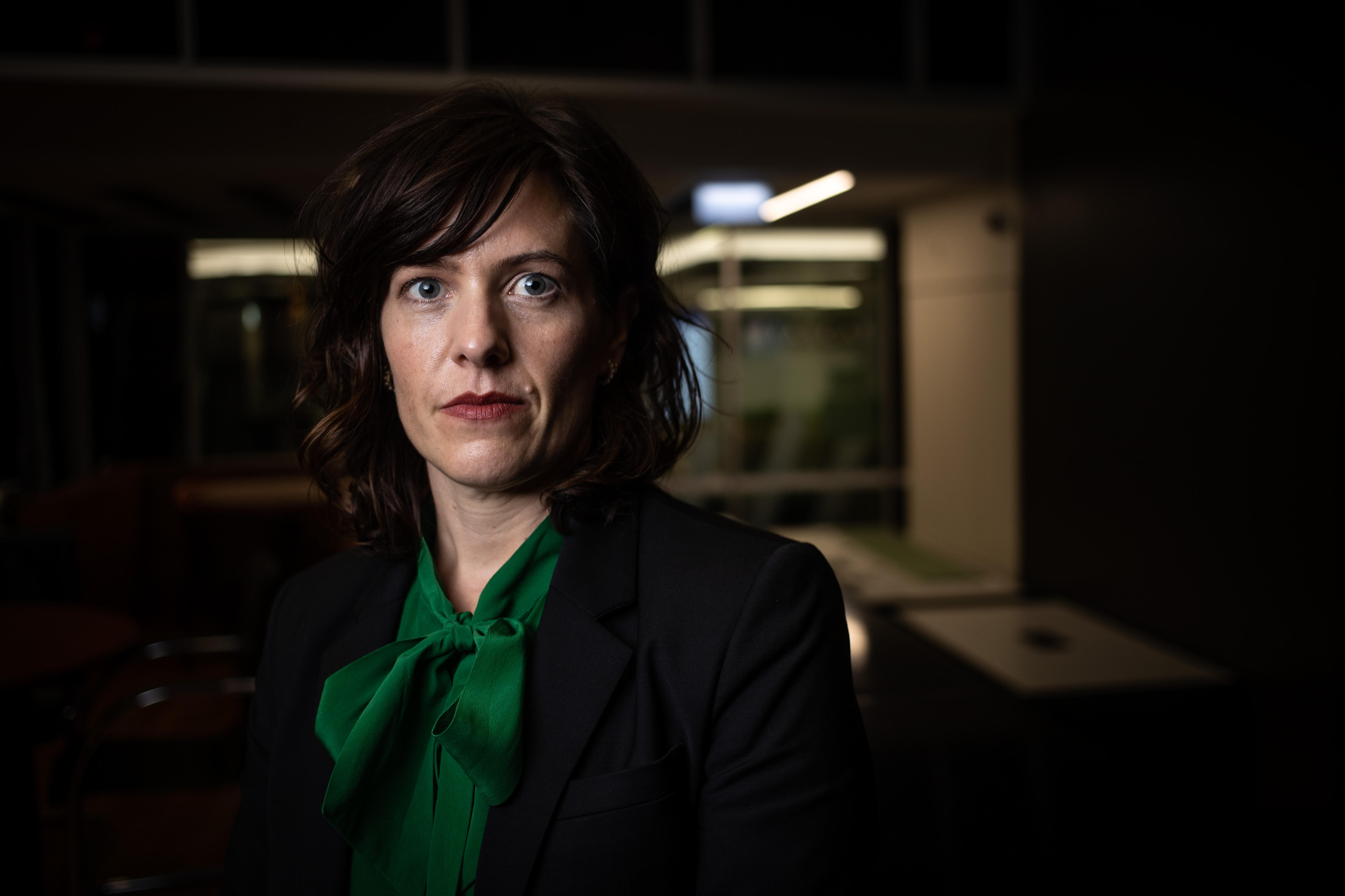 A woman in a black jacket looks at the camera with a serious expression.