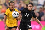 A Matildas footballer and a New Zealand player jostle each other for position as the ball bounces in front of them.